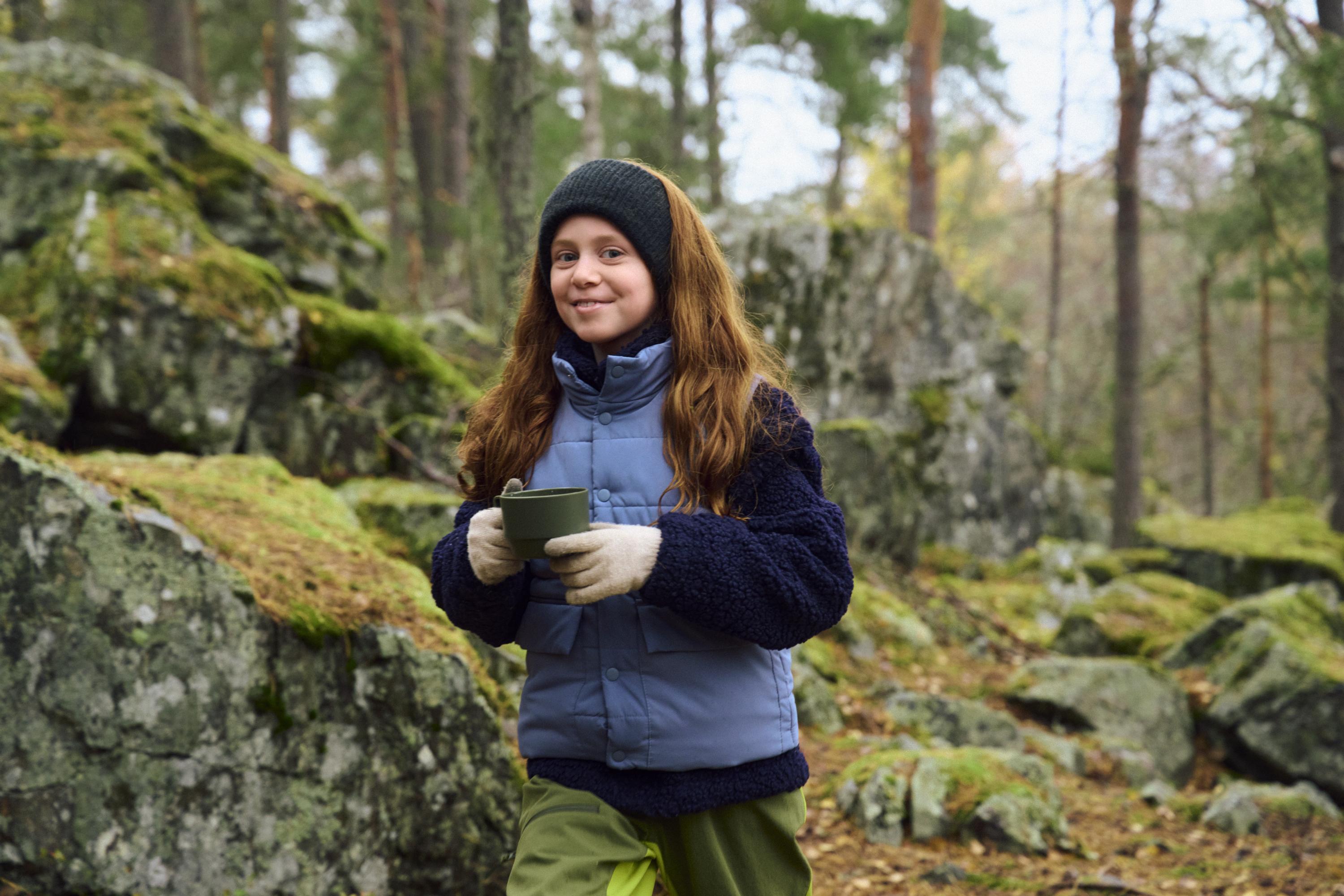 En ung tjej står i en skog och håller en mugg, omgiven av stenar och mossa.